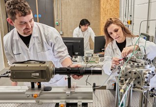 Three researchers wearing white lab coats, working with machinery and digital screens in the University of 91 Advanced Engineering Centre